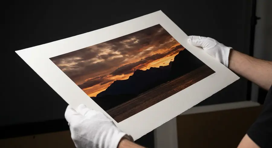 Gloved hands carefully holding a large fine art photography print showing a dramatic mountain sunset over water, professional printing quality, dark moody scene