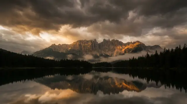 Dramatic moody mountain landscape with dark stormy sky, golden amber light on jagged rocky peaks, still reflective lake below, dark forest silhouettes, cinematic photography